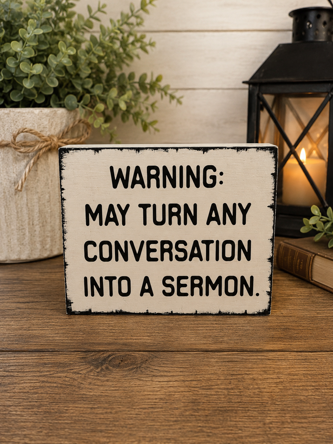 A rectangular wooden sign with a distressed cream background and black text reading 'WARNING: MAY TURN ANY CONVERSATION INTO A SERMON.' It sits on a wooden surface with a potted plant, candle lantern, and book in a styled indoor setting.