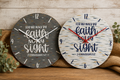 Two round decorative wall clocks with Bible verse text, displayed side by side on a rustic wooden surface with burlap and greenery.