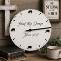 A round distressed white wall clock with black text reading 'Feed My Sheep John 21:17' and black sheep silhouettes marking the hours, styled on a rustic wooden surface with a Bible, ceramic cup, greenery, and a framed scripture print.